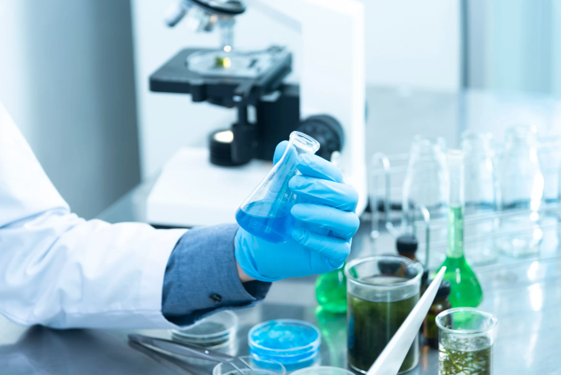 Gloved researcher holding a flask beside laboratory glassware and a microscope